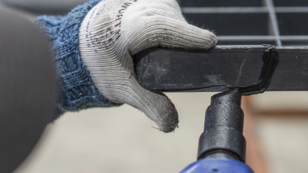 A hand wearing a glove holds a black object while using a power tool to tighten or secure it.