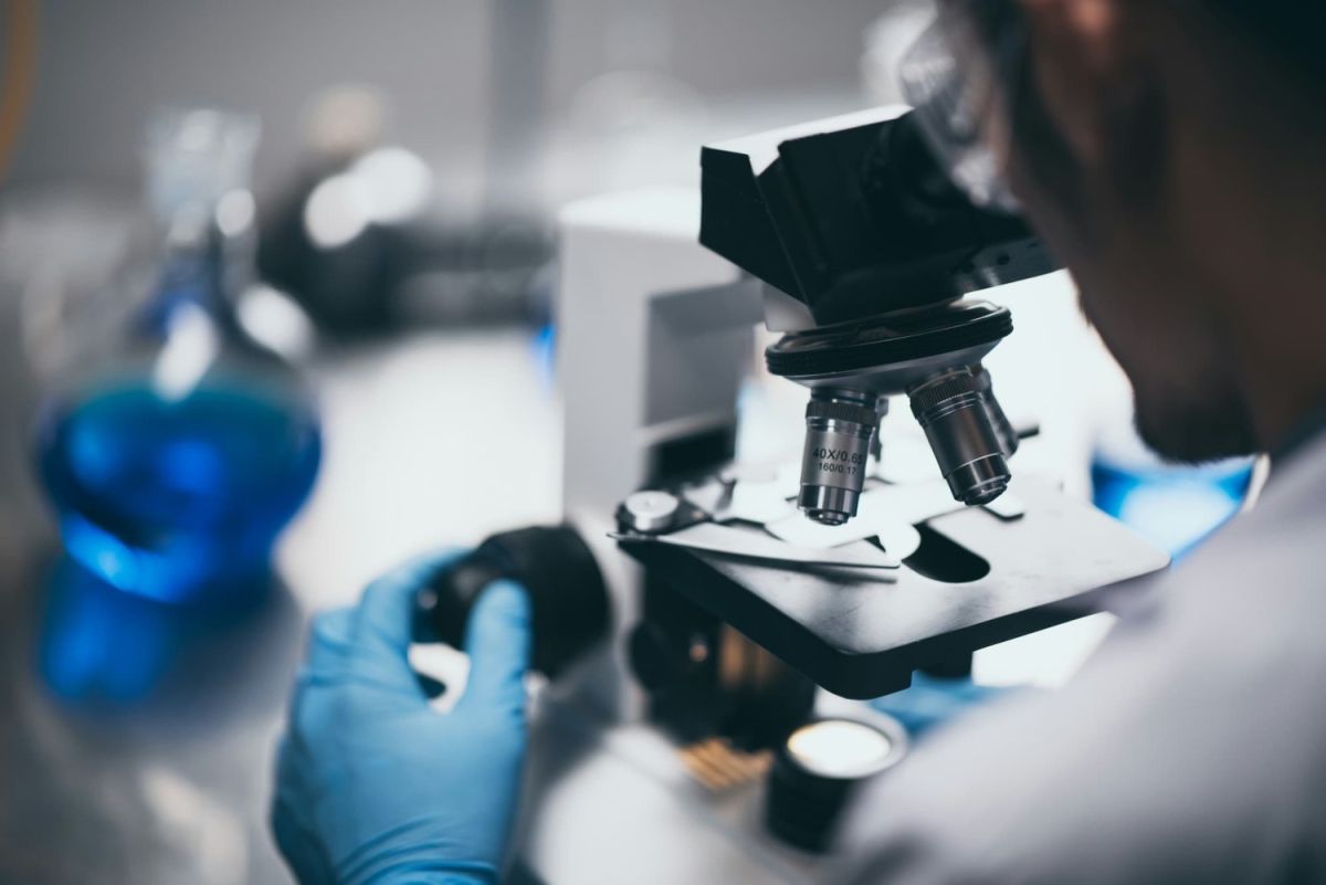 A close-up of a person in blue gloves operating a microscope in a laboratory, with blue liquid in flasks in the background.