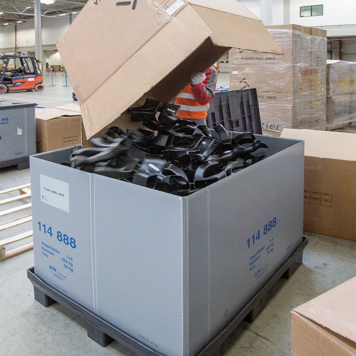 A warehouse worker in a safety vest is dumping a cardboard box filled with black plastic components into a large grey container, surrounded by pallets and other boxes in a busy storage area.