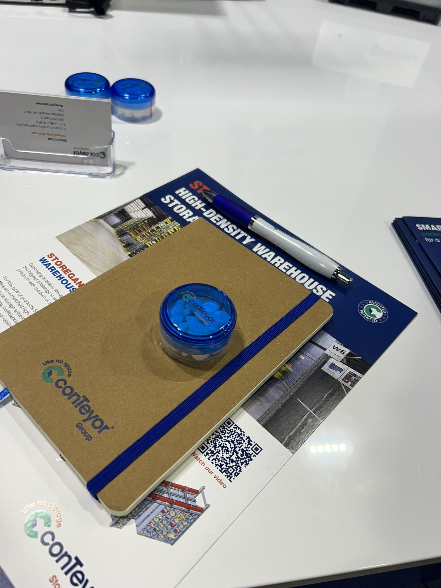 A notepad with a blue band, a blue container with white contents, a pen, and promotional materials on a white table.