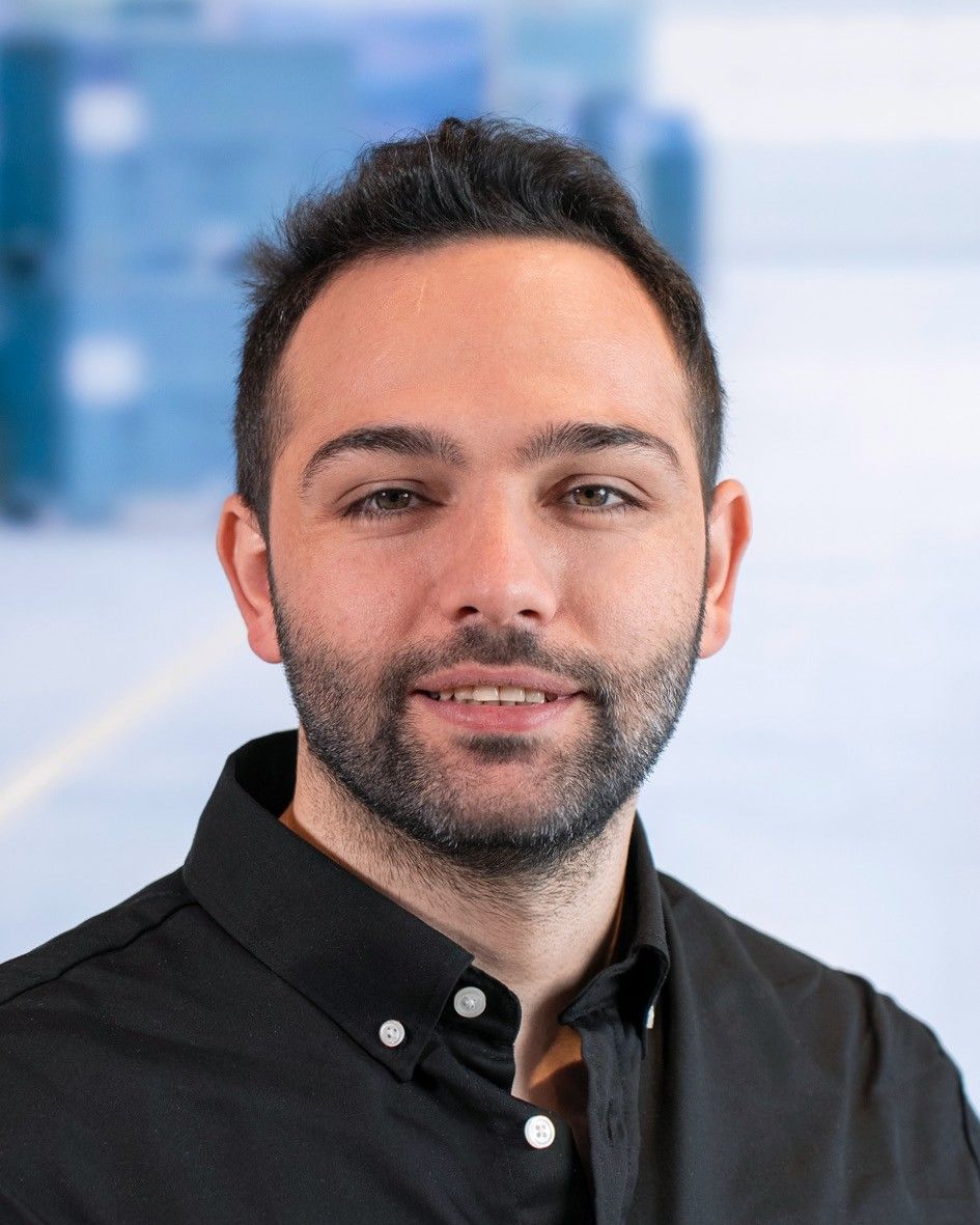 A man with a short beard and dark hair, wearing a black button-up shirt, smiles in front of a blurred background.