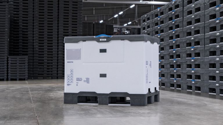 A large white reusable container with a gray lid, positioned on a concrete floor surrounded by stacked black plastic crates in a warehouse setting.
