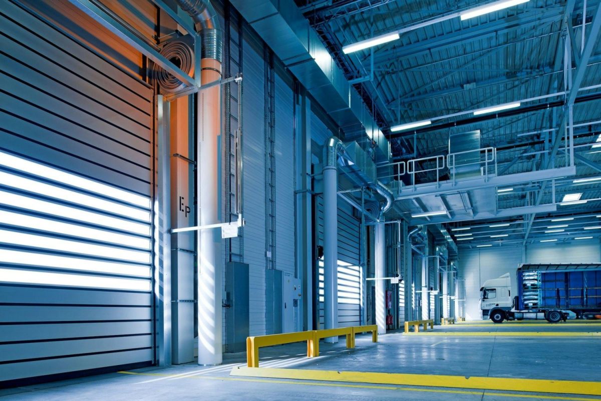 A spacious industrial warehouse interior with metallic shelving, bright overhead lights, and a yellow safety barrier, featuring a parked truck in the loading area.