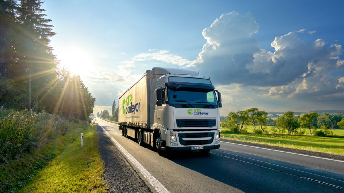 A large white truck with a green and blue logo traveling on a road surrounded by trees and fields under a bright sky.