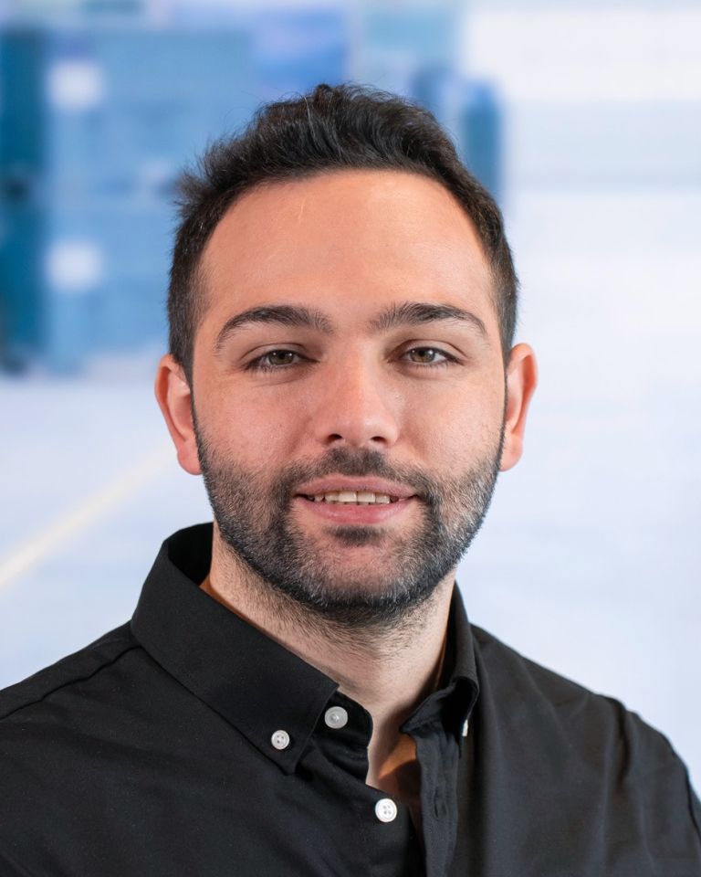 A man with a short beard and dark hair, wearing a black button-up shirt, smiles in front of a blurred background.