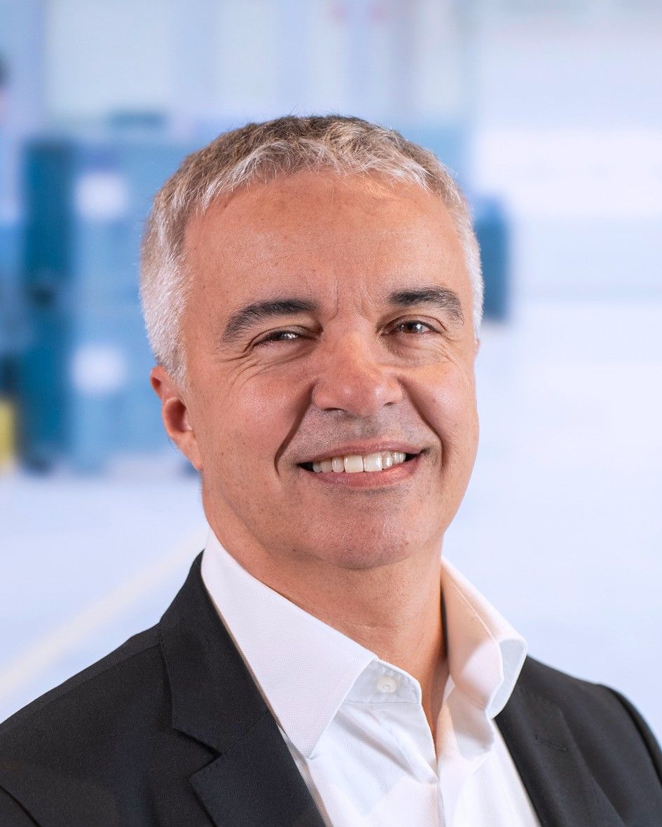 A man with short gray hair and a friendly smile, wearing a black suit and white shirt, stands in a bright space with blurred background elements.