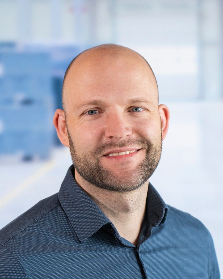A smiling man with a bald head and a short beard wearing a blue button-up shirt, set against a blurred background.
