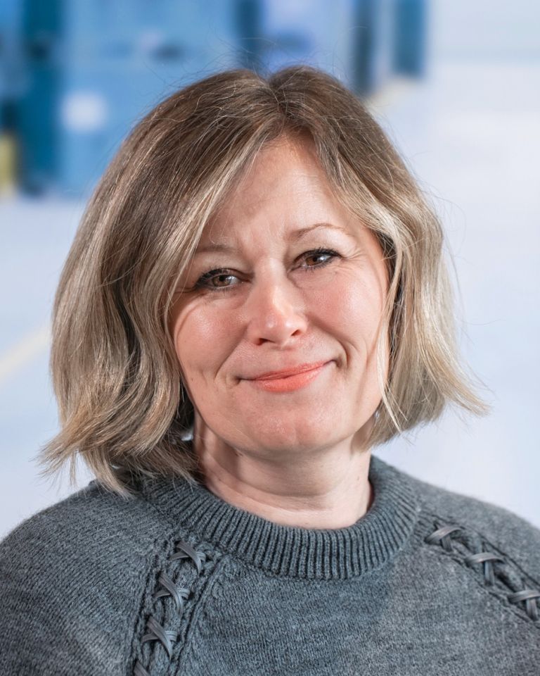 A woman with shoulder-length blonde hair, wearing a gray sweater, smiles in front of a blurred background.