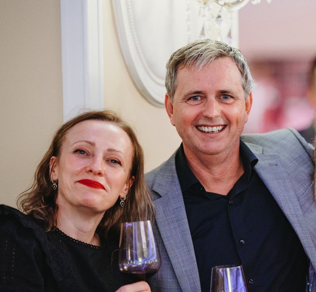 A smiling woman with shoulder-length hair, wearing a black top and red lipstick, holds a wine glass next to a smiling man in a gray blazer, both posing together against a softly lit background.