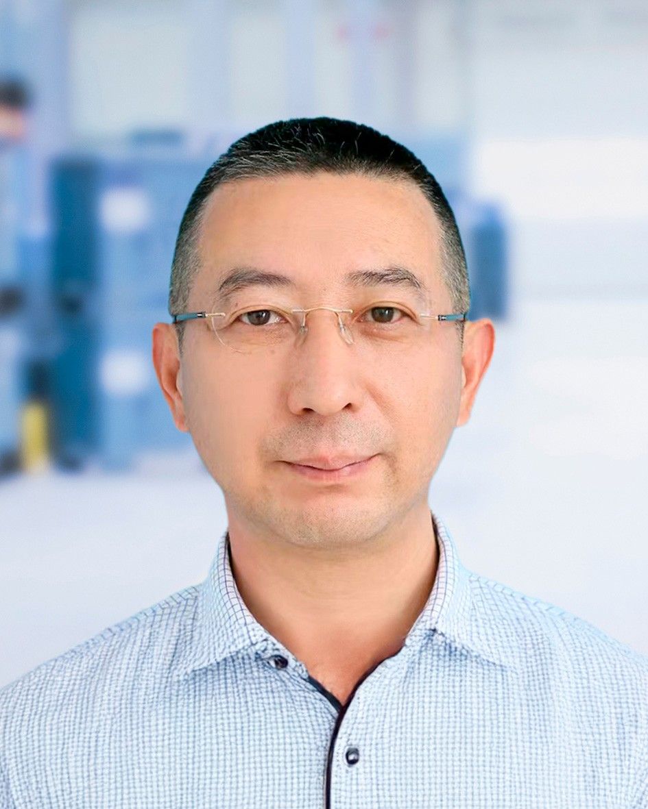 A close-up portrait of a man wearing glasses and a checkered shirt, set against a blurred background resembling an industrial or office environment.
