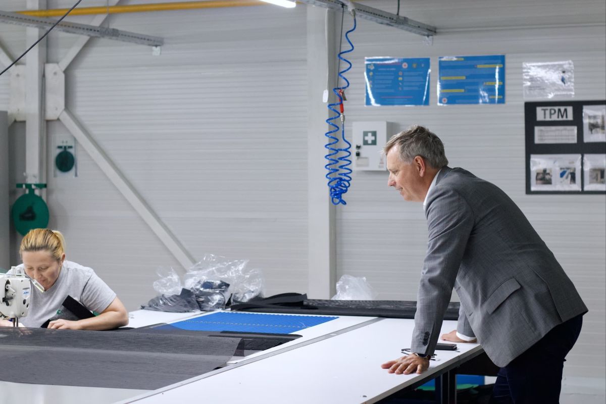 A woman operates a sewing machine while a man observes and leans against a table in a workshop environment.
