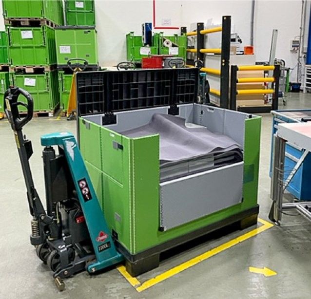 A green industrial container with a partially open lid, positioned near a pallet jack, in a warehouse setting with other green containers visible in the background.