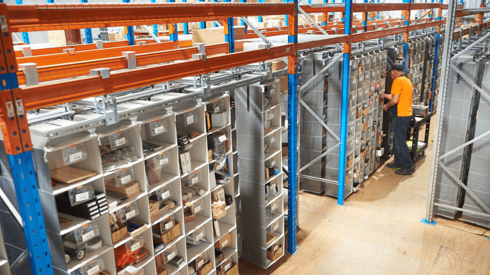 A warehouse interior with organized shelving units filled with various items, and a person in an orange shirt sorting through the shelves.