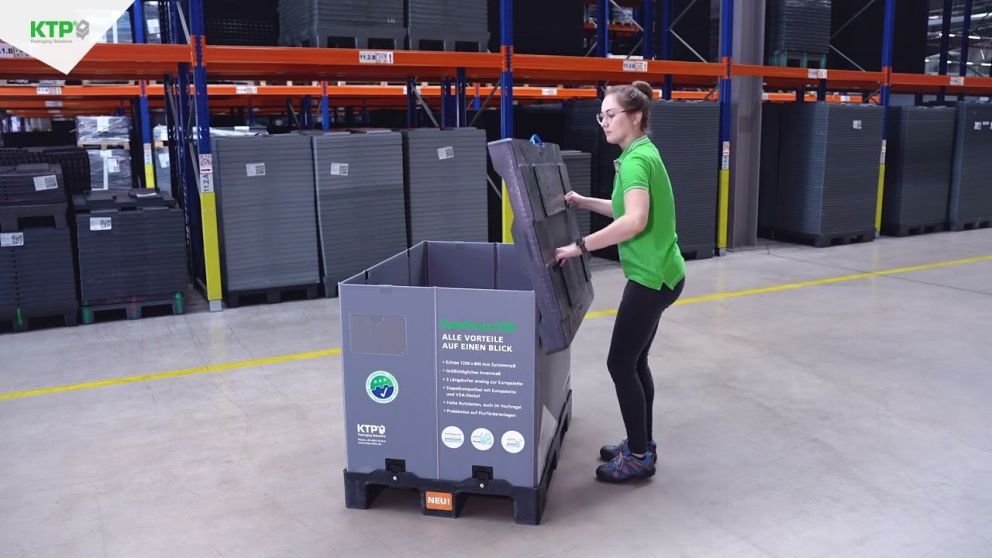 A person in a green shirt placing a lid on a large gray storage container in a warehouse filled with stacked containers.