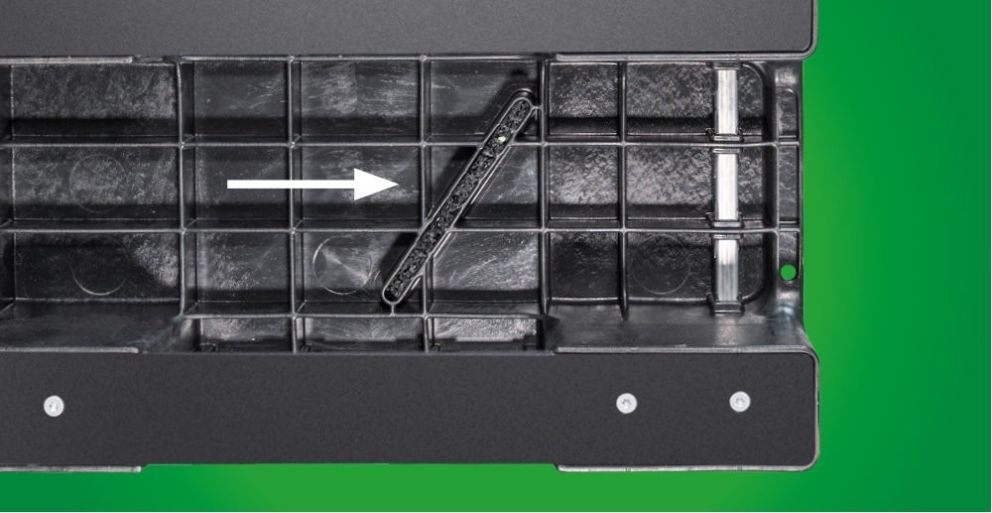 A close-up image of a black compartment with a partially inserted black tool or piece, accompanied by an arrow pointing to the right, against a green background.