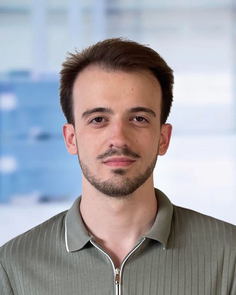 A young man with short brown hair and a light beard is looking directly at the camera, wearing a green zip-up shirt with a ribbed texture, set against a blurred background.