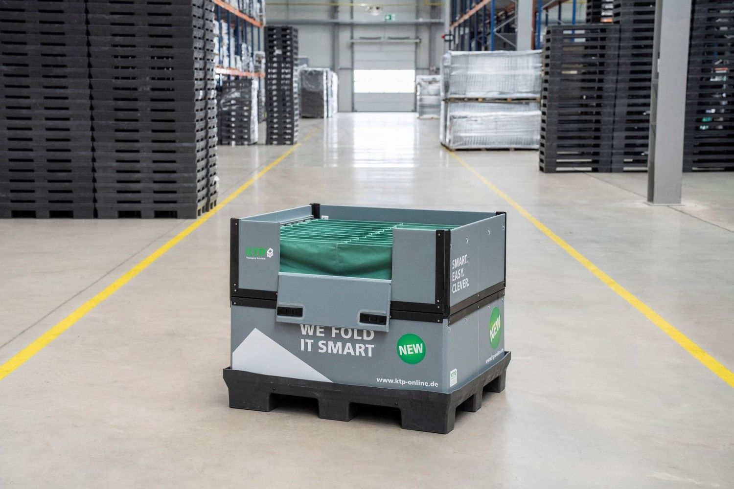 A foldable storage container with a green cover, labeled with the text "WE FOLD IT SMART," is placed in a well-lit warehouse aisle, surrounded by stacks of black pallets.