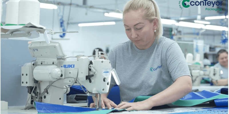 A woman working at a sewing machine, stitching blue and green materials in a factory setting.