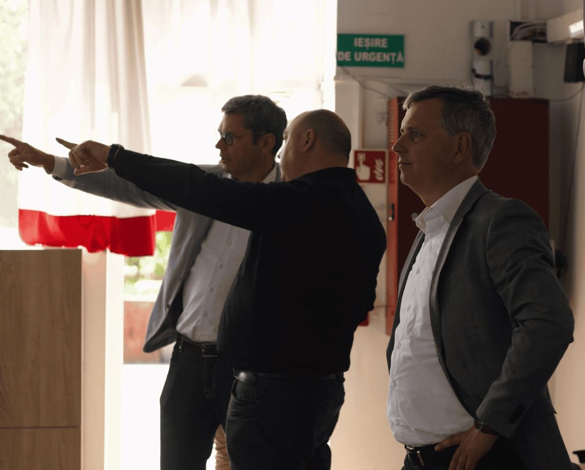 Three men standing indoors, two pointing at something off-screen while the third observes, with a red and white curtain in the background.