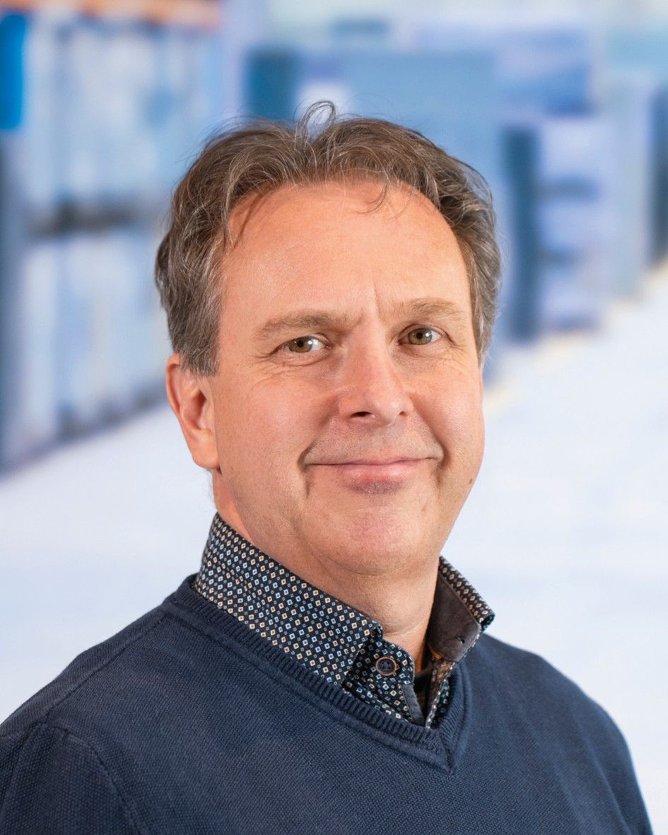 A man with short, wavy hair wearing a navy blue sweater over a patterned shirt, smiling against a blurred background.