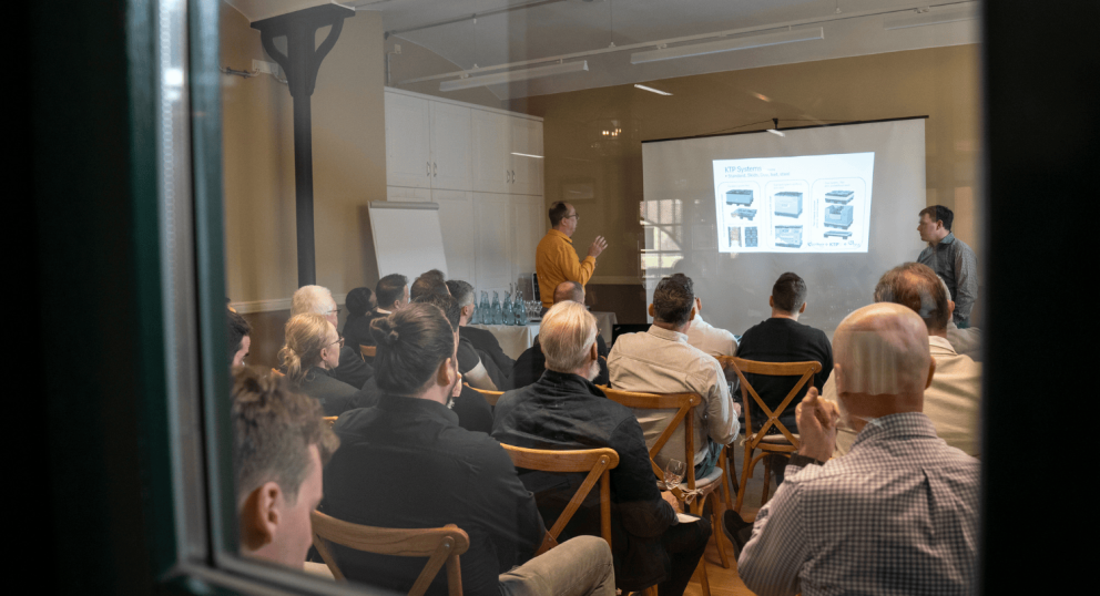 A group of people seated in a conference room attending a presentation, with one person presenting in front of a screen displaying images and text.