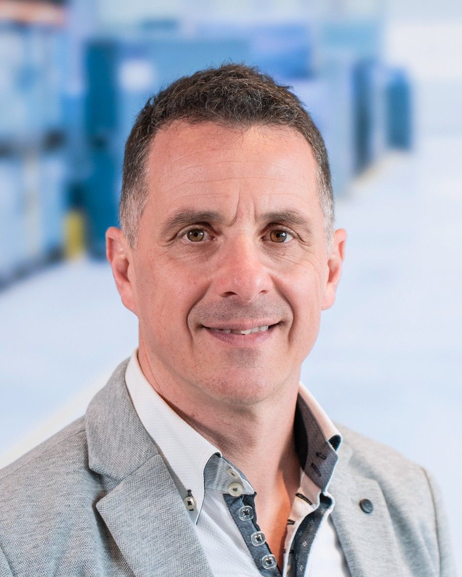 A smiling man with short dark hair, wearing a grey blazer over a light shirt, is photographed in a brightly lit environment with a blurred background.