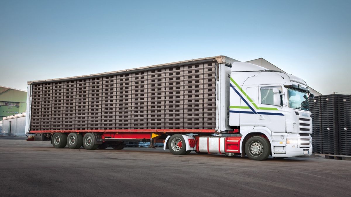 A large white semi-truck with a trailer loaded with pallets, parked in a warehouse area with a clear blue sky in the background.