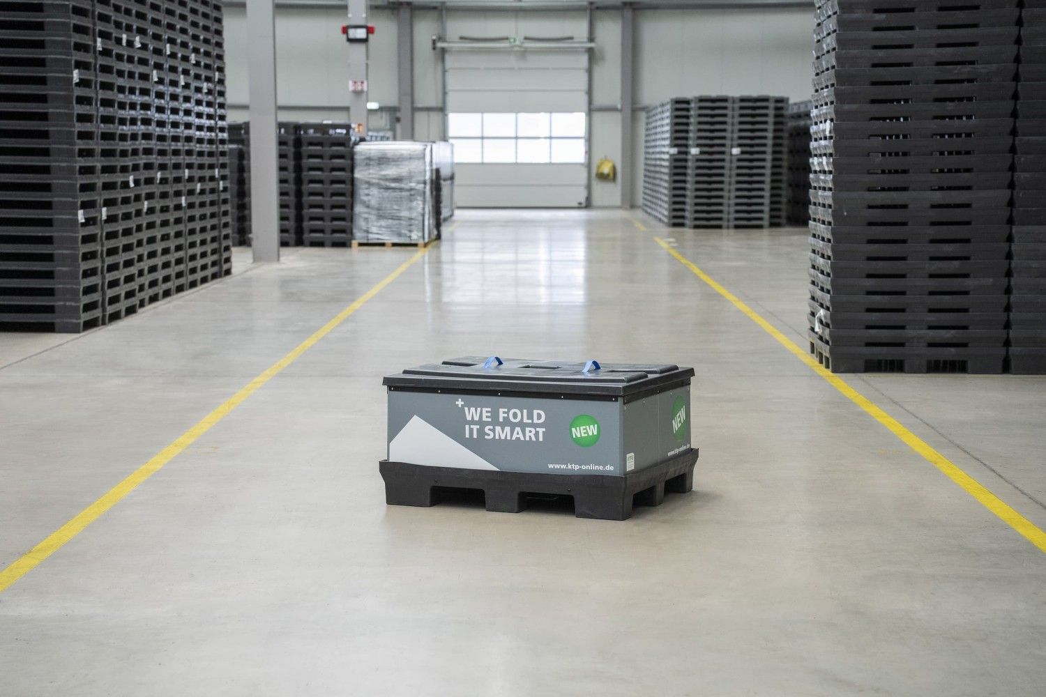 A storage box labeled "WE FOLD IT SMART" on a concrete floor in a warehouse, surrounded by stacks of pallets.