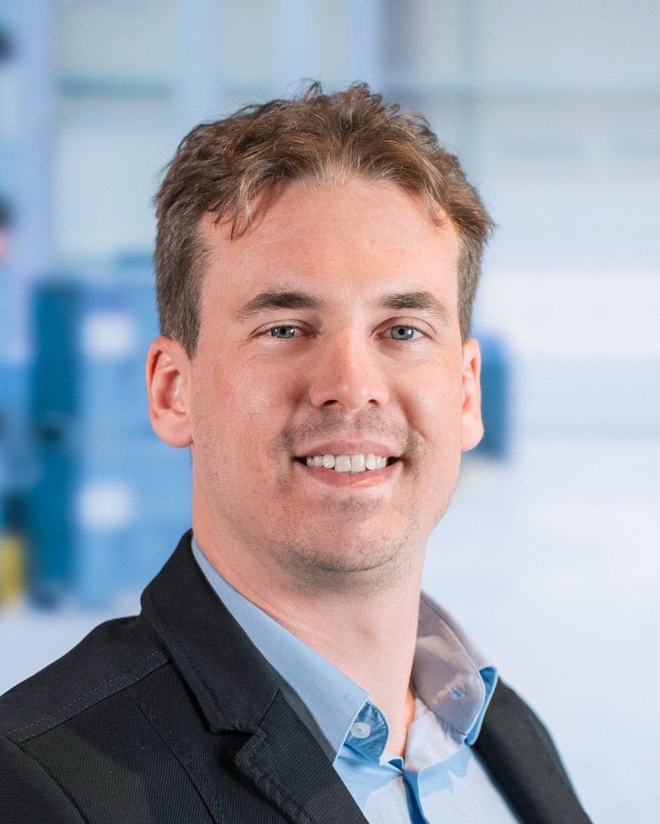 A man with short, light brown hair is smiling while wearing a black blazer over a light blue shirt, set against a blurred blue and white background.