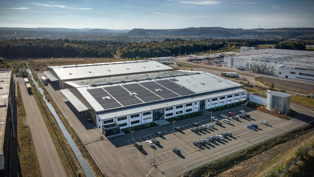Aerial view of a modern industrial building with solar panels on the roof, surrounded by a parking lot and greenery, against a background of hills and a clear blue sky.