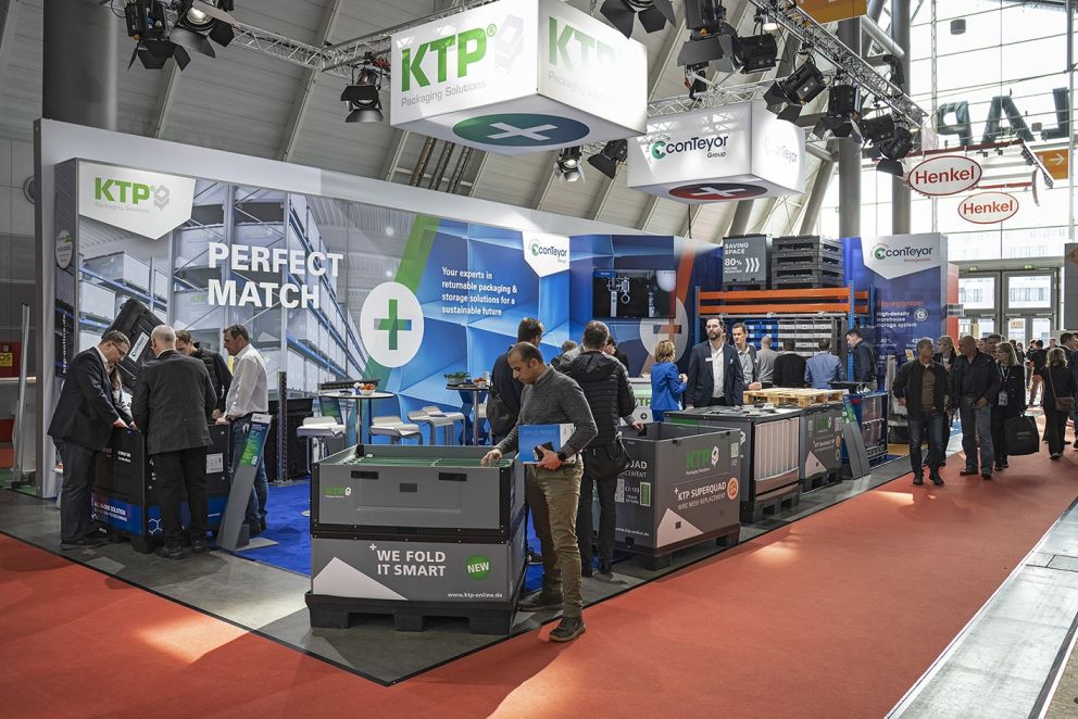 A busy exhibition booth featuring KTP packaging solutions, with visitors interacting at tables and display cases highlighting returnable packaging products, set in a modern trade show environment.
