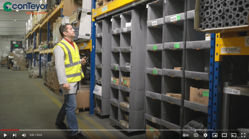 A person wearing a high-visibility vest walking through a warehouse aisle with shelves containing various boxes and bins.