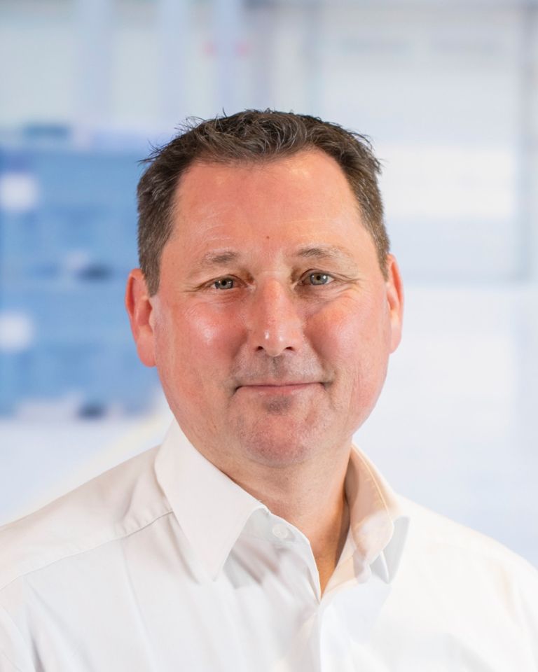 A middle-aged man with short dark hair and light facial stubble, wearing a white collared shirt, smiles at the camera against a blurred background.