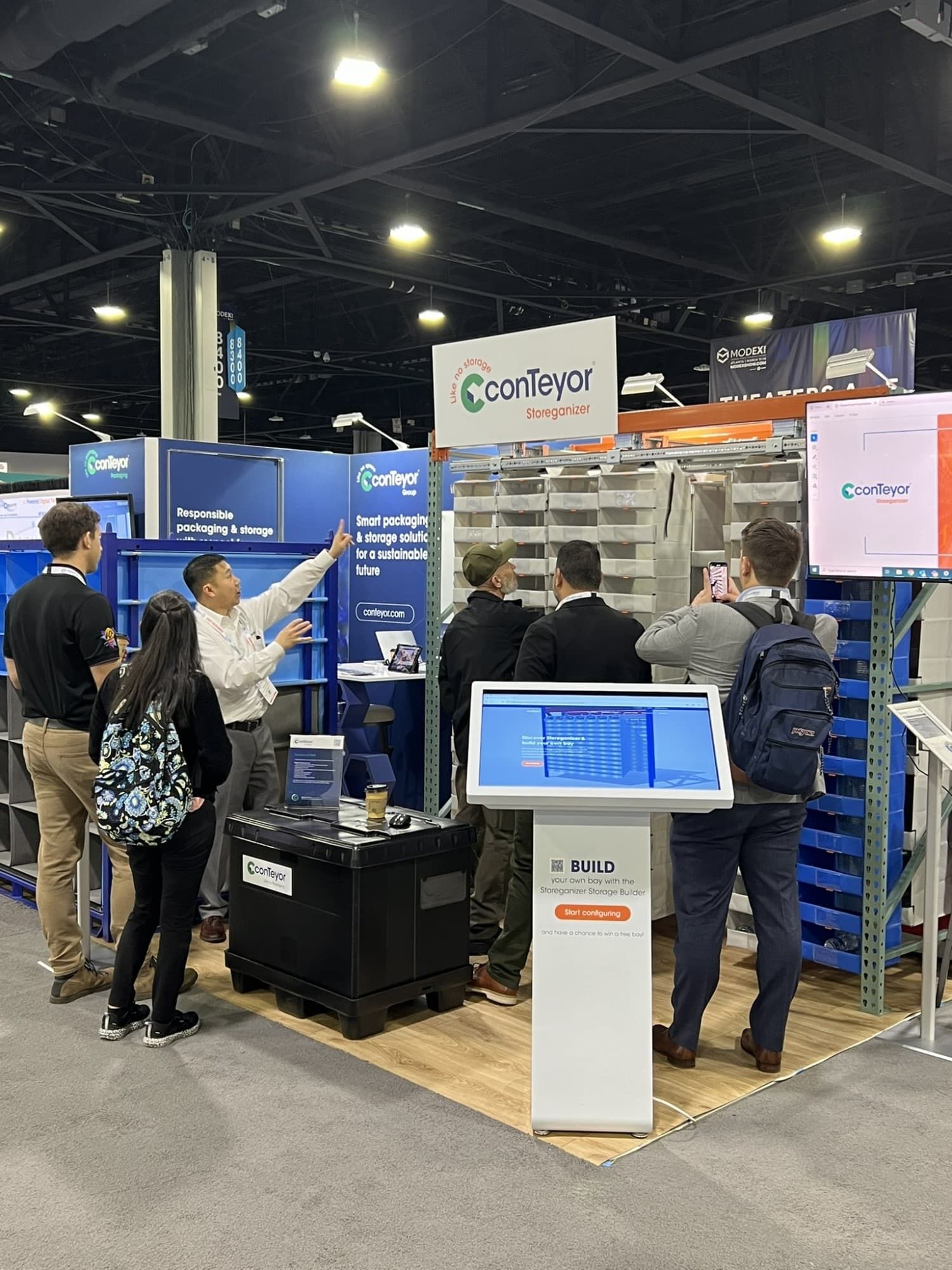 A trade show booth featuring a display of storage solutions by conTeyor, with several attendees engaged in discussion and demonstration, alongside digital screens and promotional signage.