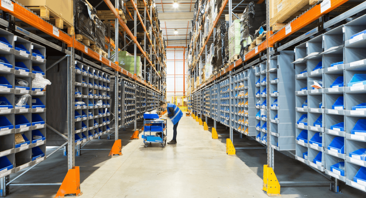 A person in a blue safety vest organizing items on a cart in a large, well-lit warehouse aisle filled with shelving units and storage bins.