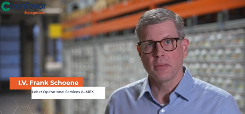 A man with glasses and short hair speaks in a warehouse setting with shelves in the background, displaying various items; text in the image identifies him as I.V. Frank Schoene, Leiter Operational Services ALMEX.