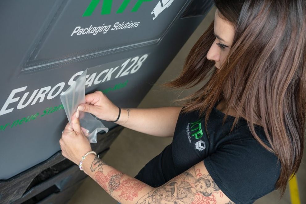 A woman with long hair and tattoos is applying a clear plastic sheet to a surface featuring the text "EuroStack 128" as part of her work at a packaging solutions company.