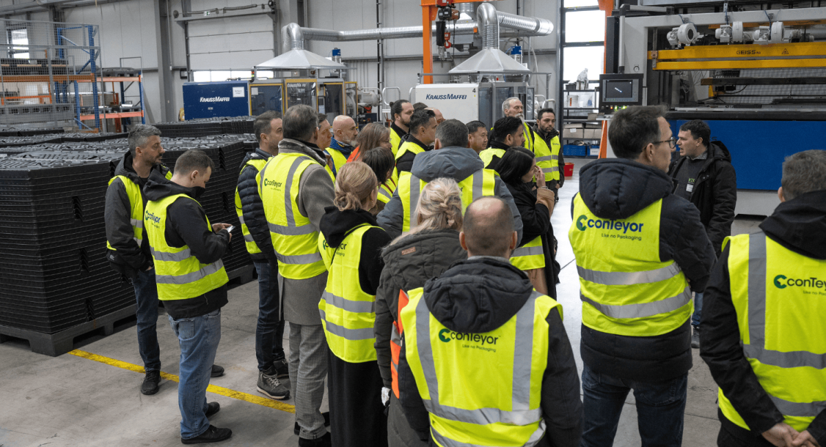 A group of people wearing yellow safety vests and standing in a factory space, discussing or listening to a presentation near machinery.
