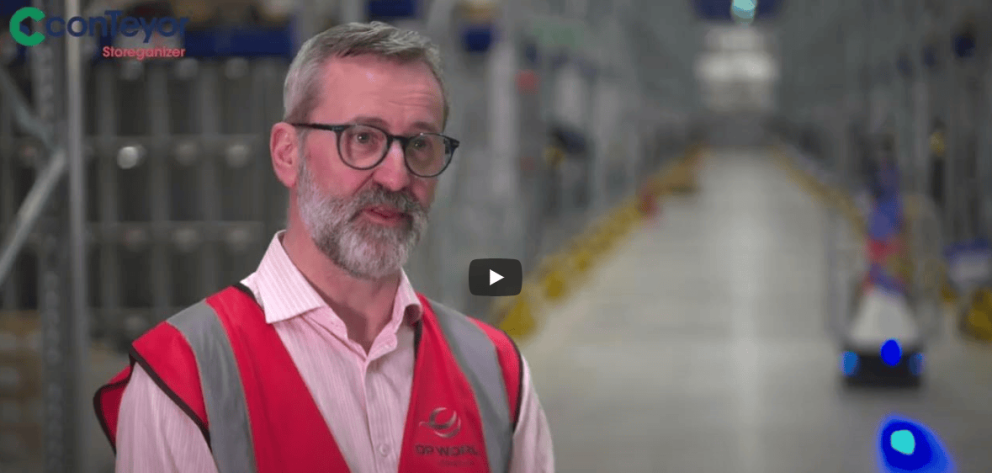 A man with glasses and a beard wearing a red work vest speaks in a warehouse setting, with shelves and automated robots visible in the background.