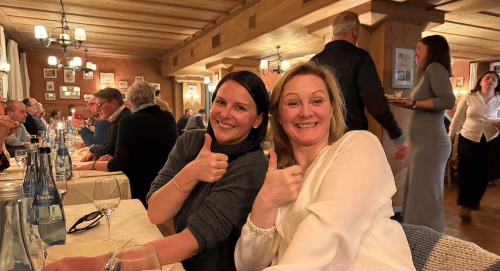 Two women sitting at a dinner table giving thumbs up, surrounded by other people and a cozy restaurant atmosphere.
