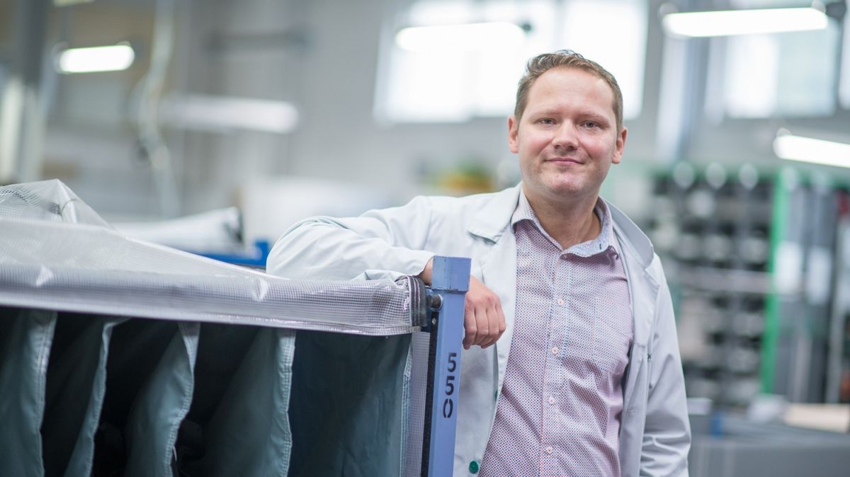 A man in a lab coat is standing next to a large piece of equipment in a bright industrial setting.
