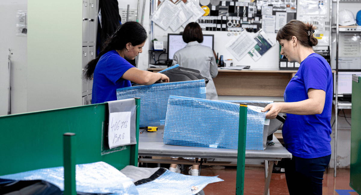 Two women in blue shirts are working at a table in a workshop, assembling or organizing items within a blue mesh container, while a third person is seen in the background at a computer.