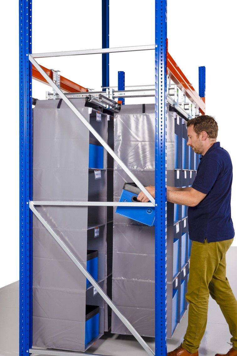 A person reaching into a storage shelving unit with gray fabric compartments and blue boxes, while wearing a navy blue shirt and light-colored pants.
