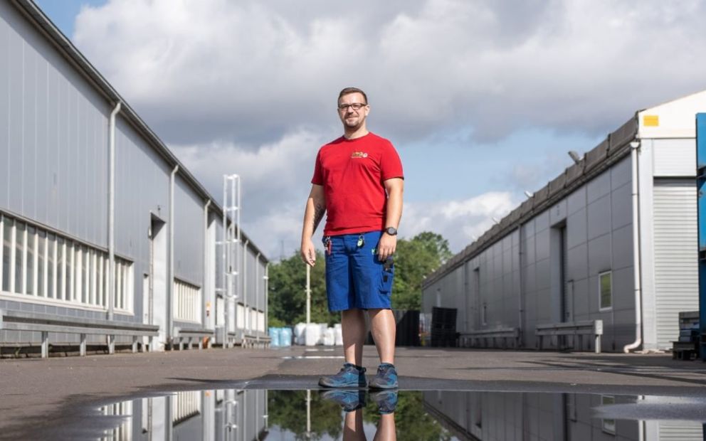A man in a red shirt and blue shorts stands on a paved area with a puddle reflecting the sky and buildings, surrounded by industrial structures and greenery.