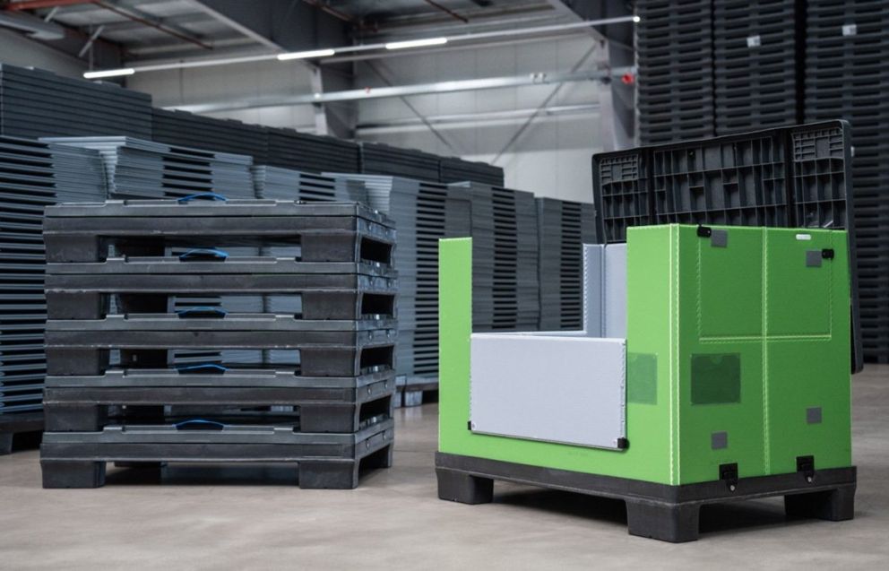 A bright green container with gray accents positioned next to a stacked pallet in a warehouse setting, with additional pallets visible in the background.