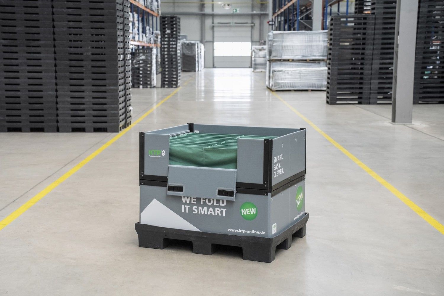 A gray and black folding container on a pallet with green interior and labels indicating "NEW," "SMART. EASY. CLEVER." in a warehouse setting with shelves of dark items in the background.