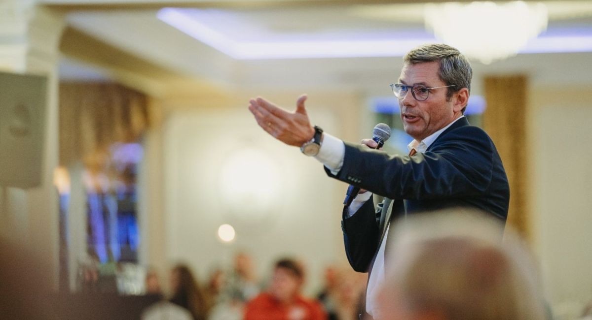 A speaker gestures while addressing an audience, holding a microphone in a formal setting.