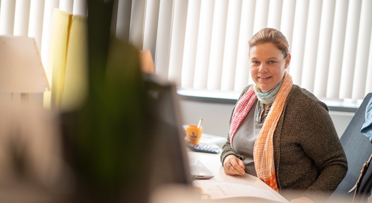 A woman sitting at a desk with a computer, smiling, wearing a scarf, in an office setting with window blinds in the background.