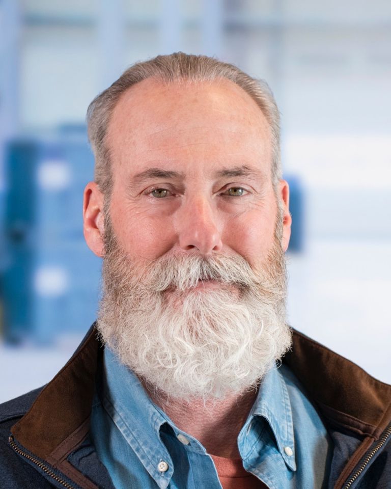 A man with a full, gray beard and short hair, wearing a blue shirt and a brown jacket, smiling against a blurred background.