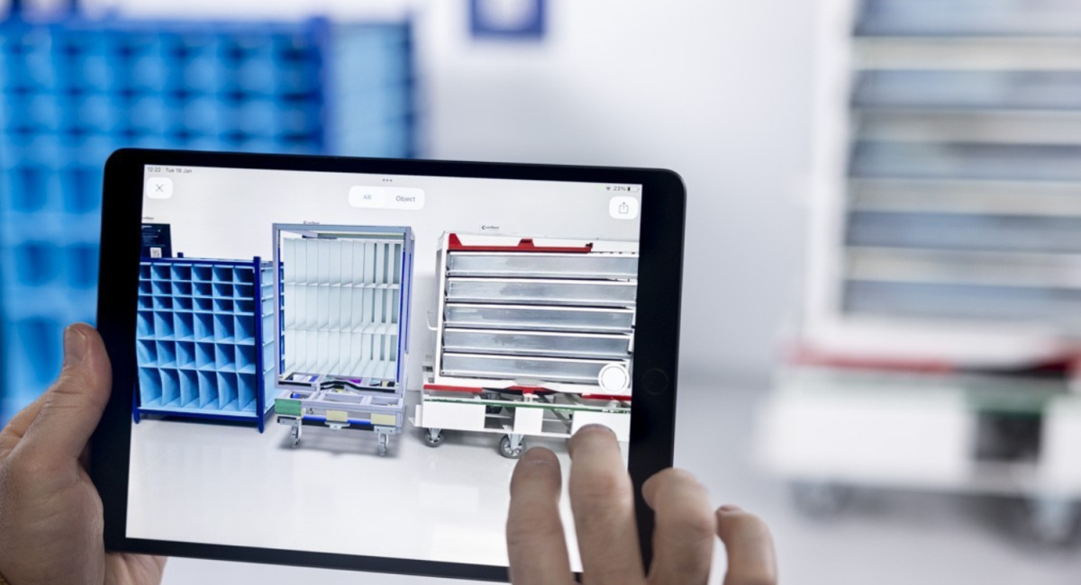 A person holds a tablet displaying a 3D model of a storage cart alongside real storage solutions in a warehouse setting, with blue storage bins in the background.